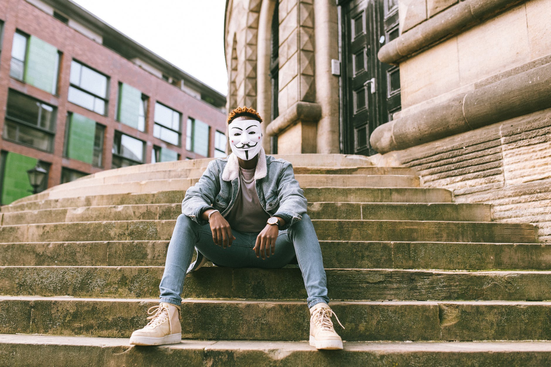 black man in anonymous mask sitting on stairs on street